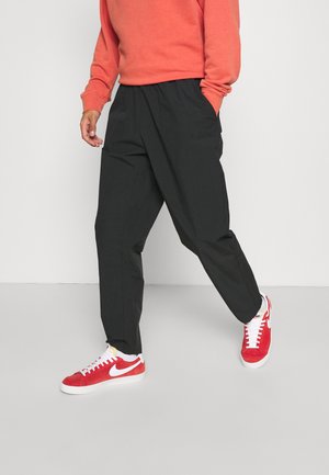Person mit lockerer schwarzer Hose, roten und weißen Nike-Sneakern und einem korallenfarbenen Langarmshirt, die vor einem schlichten Hintergrund geht.