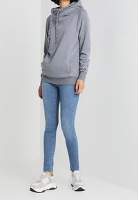 Šedá mikina s kapucí a šálovým límcem, se šňůrkami a bočními kapsami, spojená se světle modrými slim džíny a robustními teniskami.