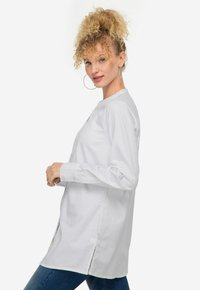 Camicia bianca a maniche lunghe con colletto rotondo, chiusura anteriore con bottoni e fessure laterali, realizzata in tessuto liscio. Abbinata a jeans blu.