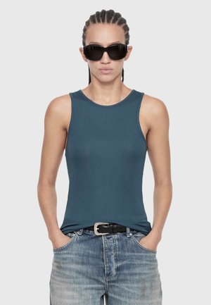 Woman with braided hair wearing large black sunglasses, a sleeveless dark teal top, blue jeans, and a black belt, hands in pockets.