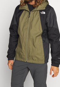 Chaqueta con capucha en verde oliva y negro, con tejido transpirable, puños elásticos y logotipo en el pecho. Diseño ligero.