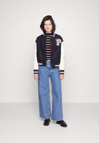 Giacca varsity navy e crema con patch a lettere, dolcevita a righe, jeans a gamba larga e scarpe nere. Design semplice con colori a contrasto.