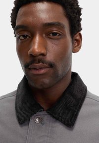 Grå jakke med en sort fløjlskrave, trykknaplukning og en subtil tekstur, tæt indrammet omkring ansigtet på en model.