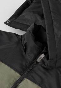 Gros plan sur le col d'une veste noire et vert olive avec fermeture éclair et détails de bouton-pression sur un tissu matelassé.