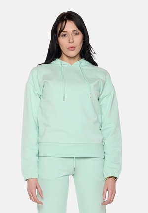 Femme aux longs cheveux foncés portant un sweat à capuche vert clair et un pantalon assorti, debout devant un fond uni.