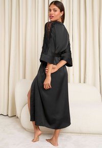 Robe en satin noir avec des accents en dentelle sur les manches, une taille ceinturée et une fente sur le côté. Texture lisse avec un design fluide.