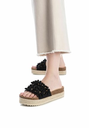Sandalias de plataforma negras con acentos florales. Presentan un material similar al ante, suela de corcho y banda de yute trenzado. El diseño de dedo abierto muestra los pies desnudos.
