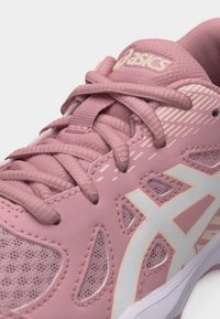 Κοντινό πλάνο ροζ αθλητικού παπουτσιού ASICS με υφασμάτινες και συνθετικές επικαλύψεις, λεπτομερείς ραφές, διακοσμητικό δίχτυ και κορδόνια που σχηματίζουν σταυρό.