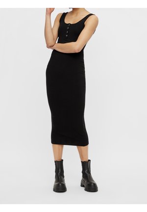 Femme portant une robe midi noire sans manches avec des boutons et des bottines noires épaisses, debout les bras croisés.