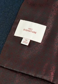 Bordeauxfarbener Stoff mit geometrischem Muster, Etikett mit "NEXT SIGNATURE", Größe 40L, in Vietnam hergestellt, vor einem strukturierten blauen Hintergrund.