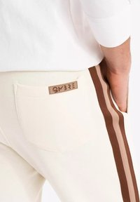 Pantalones joggers crema con rayas laterales marrones, con un bolsillo trasero y una etiqueta rectangular "OMBRE". Textura suave y lisa.