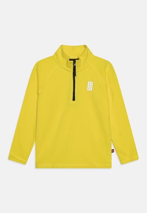 Gelber Langarm-Pullover mit schwarzem Halbreißverschluss und kleinem weißem rechteckigem Aufnäher mit sechs gelben Punkten auf der Brust.