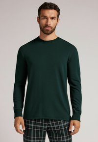 Ciemnozielona koszula z długim rękawem, wykonana z miękkiego materiału, z okrągłym dekoltem i dopasowanymi mankietami, zestawiona z piżamowymi spodniami w kratkę.