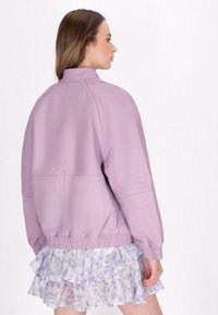 Pastel paarse bomberjack gemaakt van gestructureerd materiaal met een relaxte pasvorm, voorzien van elastische manchetten en een geribde tailleband. Rimpelrok zichtbaar eronder.