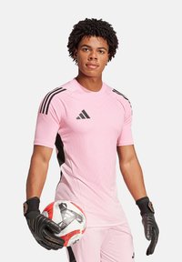 Maillot de gardien de but rose avec des accents noirs et trois rayures verticales noires sur les épaules. Gants noirs et un ballon de football blanc et rouge.