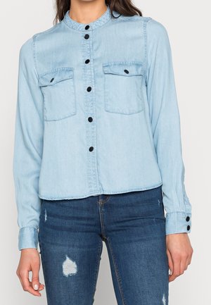 Lyseblå denim-skjorte med knapper og to frontlommer med klap, båret med mørkeblå slidte jeans på en stående person.