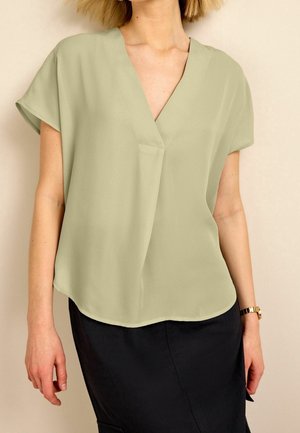 Blouse vert clair à manches courtes avec un décolleté en V et une coupe ample, fabriquée en tissu lisse ; portée sur une jupe sombre. Présente une petite poche avant.