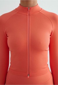 Kinnitatud sporttop erksas korallis. Valmistatud siledast, venivast kangast, millel on sooniktekstuur. Omab kõrget kraed ja sobivat lõiget.