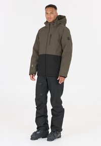 Zweifarbige Jacke mit olivgrünem Oberteil und schwarzem Unterteil, ausgestattet mit Kapuze, Frontreißverschluss und einer Brusttasche. Getragen mit schwarzen Skihosen und Stiefeln.