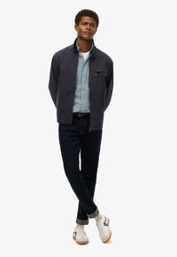 Marineblå jakke over en lyseblå stribet skjorte, mørke jeans, hvide sneakers med grå detaljer, stående pose mod en hvid baggrund.