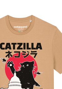 Beige katoenen t-shirt met een ronde halslijn, afgebeeld met een grafische weergave van een zwarte kat, een rode zon, een wit gebouw en illustraties van een helikopter. De tekst luidt "CATZILLA."