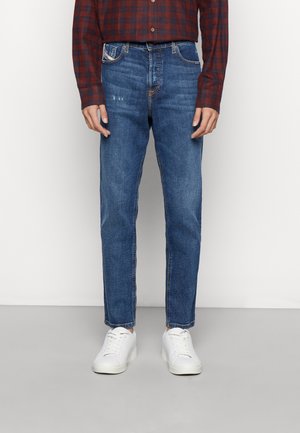 Mand iført blå slim-fit jeans, hvide sneakers og en rød-svart ternet langærmet skjorte, stående foran en ensfarvet baggrund.