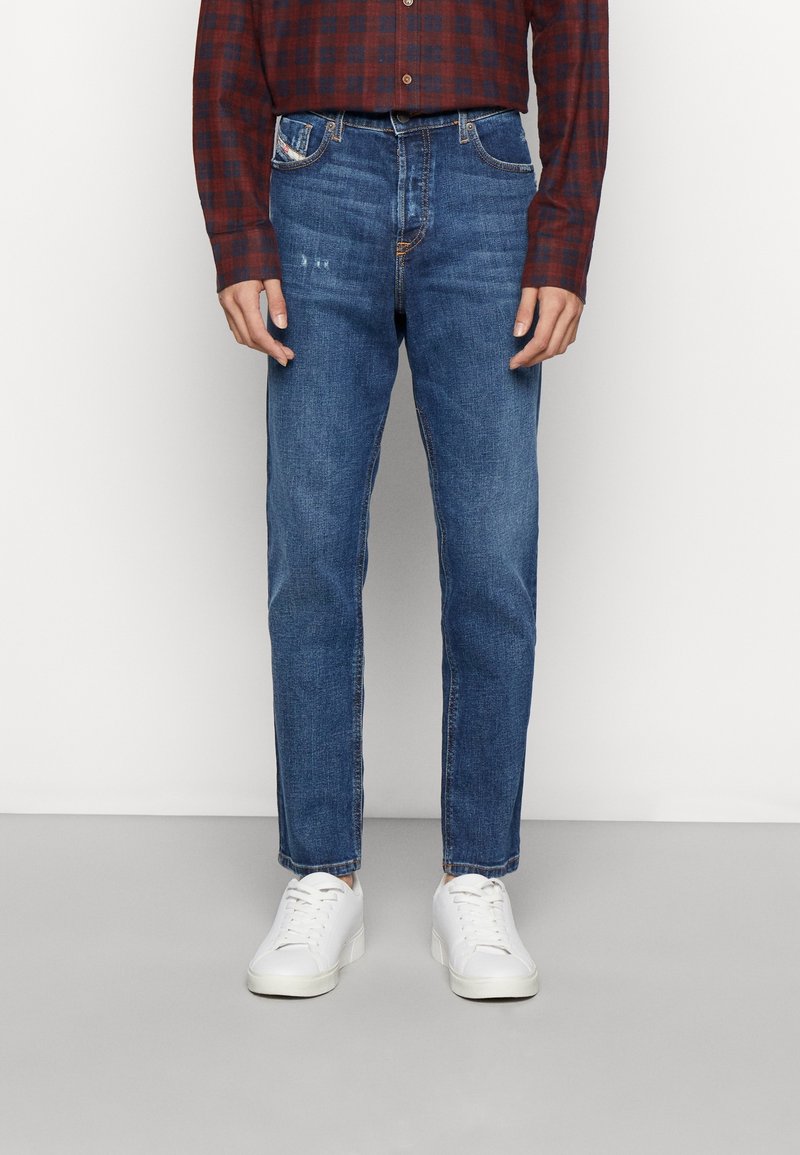 Homme portant un jean slim bleu, des baskets blanches et une chemise à carreaux rouge et noir à manches longues, debout devant un fond uni.