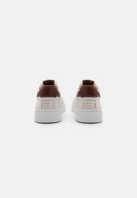 Baskets en cuir blanc avec des accents en cuir marron au talon, semelle texturée et un patch logo à l'arrière. Design minimaliste et lignes épurées.