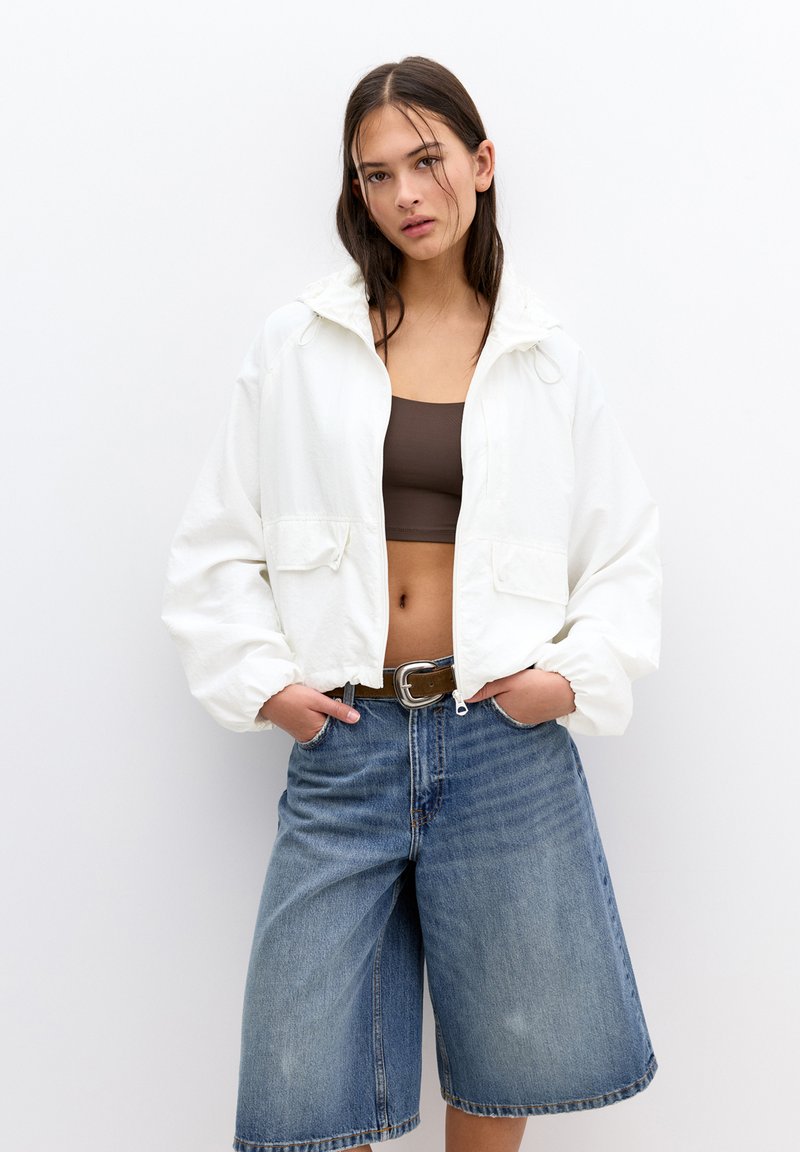 Veste blanche courte avec capuche, deux poches à l'avant, associée à un haut marron et un short en jean bleu. Le tissu a une texture lisse.