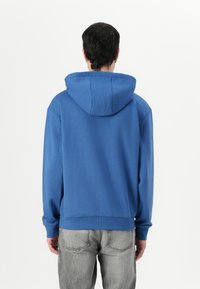 Modra bombažna kapuca s prostorno kapuco, rebrastimi manšetami in robom ter gladko teksturo, oblečena z lahkimi sivo-denim hlačami.