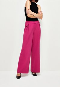 Fuchsiové kalhoty širokého střihu s páskem ve stylu papírového pytle a detailem mašle, spárované s černým bezrukávovým topem a černými podpatky. Hladká textura.