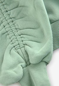 Felpa verde chiaro con dettagli arricciati elastici, tessuto testurizzato e polsini a coste. Finitura liscia con materiale morbido ed elastico.