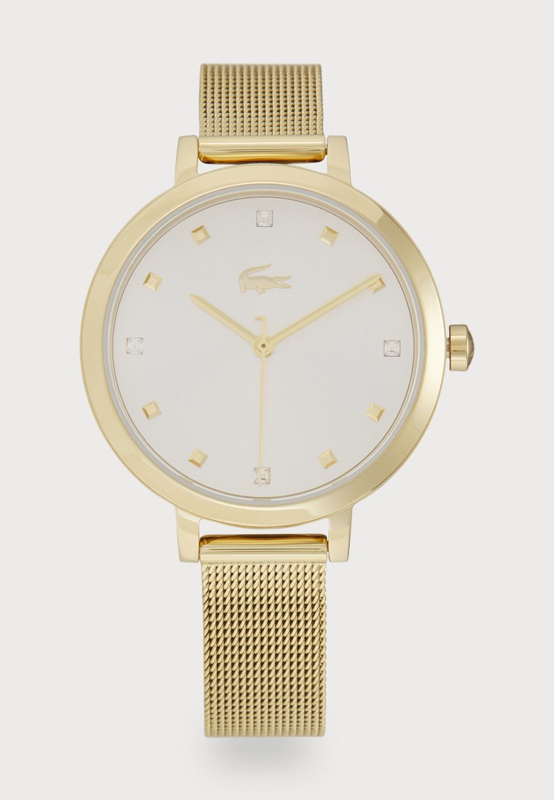 Gullur metallklokke med hvit urskive, som har kvadratiske timemarkører, minimalistisk design og nettingarmbånd. Lacoste-logoen er vist.