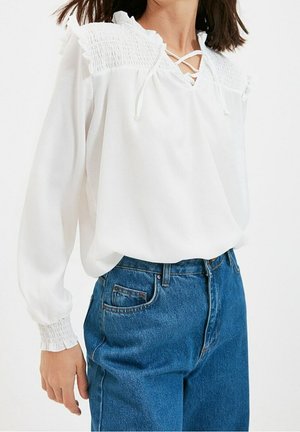 Kobieta ubrana w białą bluzkę ze ściągaczami na karczku i mankietach, w połączeniu z wysokim stanem jeansów w kolorze niebieskim na jednolitym białym tle.