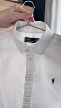 Hvit skjorte med krage laget av bomull, med en enkelt svart polo-spillerlogo på venstre bryst og frontlukking med knapper, samt en glatt tekstur.