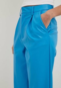 Helle blaue Kunstlederhosen mit glatter Textur, ausgestattet mit einer gefalteten Vorderseite und seitlichen Taschen. Der Bund ist gerade und strukturiert.