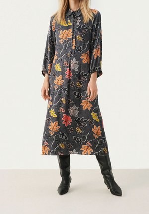 Femme portant une robe midi manches longues gris foncé avec des motifs colorés de feuilles d'automne et des bottes noires montantes jusqu'aux genoux.