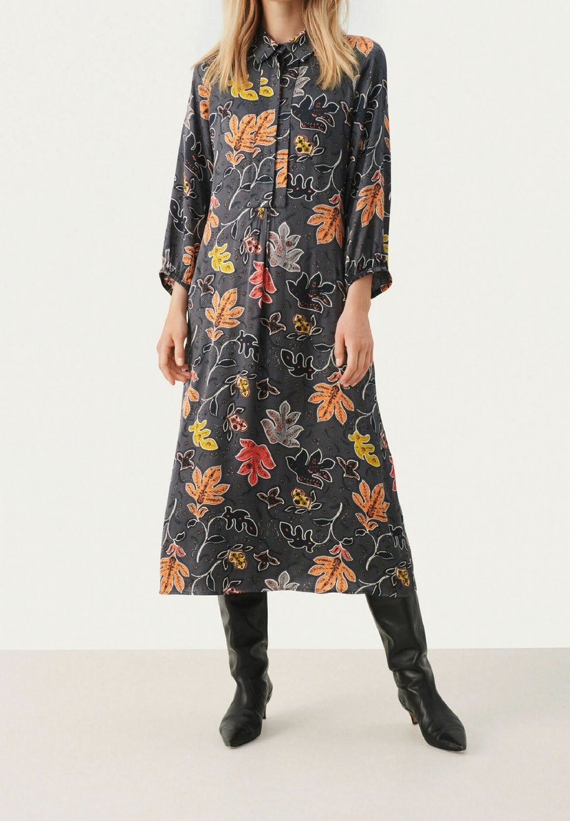 Femme portant une robe midi manches longues gris foncé avec des motifs colorés de feuilles d'automne et des bottes noires montantes jusqu'aux genoux.