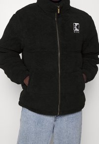 Giacca nera pelosa con zip, con le mani nelle tasche, caratterizzata da un piccolo logo bianco a forma di "K" sul petto in alto a sinistra, indossata con jeans azzurri.