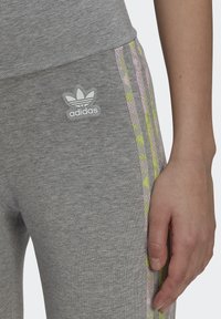 Pelēki sporta legingi ar pielāgotu dizainu, uzgūstot sudraba Adidas logo uz gurniem un krāsainām rakstainām sānu svītrām rozā un zaļā krāsā.
