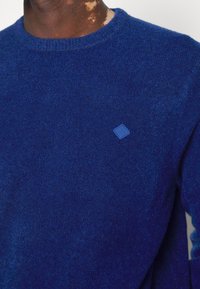 Blå stickad tröja med rund halsringning, mjuk struktur och en liten blå diamantlogotyp på bröstet.