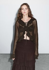 Jonge vrouw met lang golvend haar, gekleed in een doorzichtig bruin ruffles-topje dat bij de borst is vastgebonden en een gelaagde bruine rok, staande tegen een lichte muur.