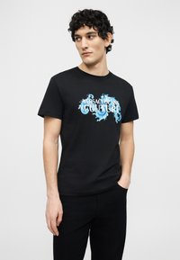 Νεαρός άνδρας με σγουρά σκούρα μαλλιά, φοράει μαύρο T-shirt με μπλε κείμενο και γραφικά "Versace Jeans Couture", με το ένα χέρι στην τσέπη, κοιτάζοντας προς τα αριστερά.