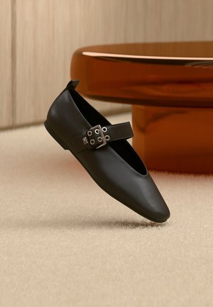Black leather flat shoe with a pointed toe and side strap, featuring silver hardware. Smooth texture against a neutral carpet background.