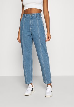 Vrouw die een blauw spijkerbroek met rechte pijpen en hoge taille draagt, een wit cropped tanktop en witte sneakers met een zwart logo.