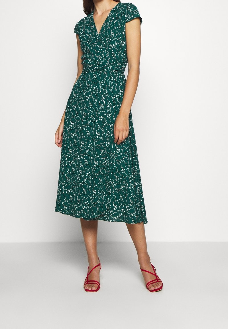 Femme portant une robe portefeuille midi verte avec imprimé floral blanc et manches courtes, associée à des sandales rouges à talons hauts et à brides.