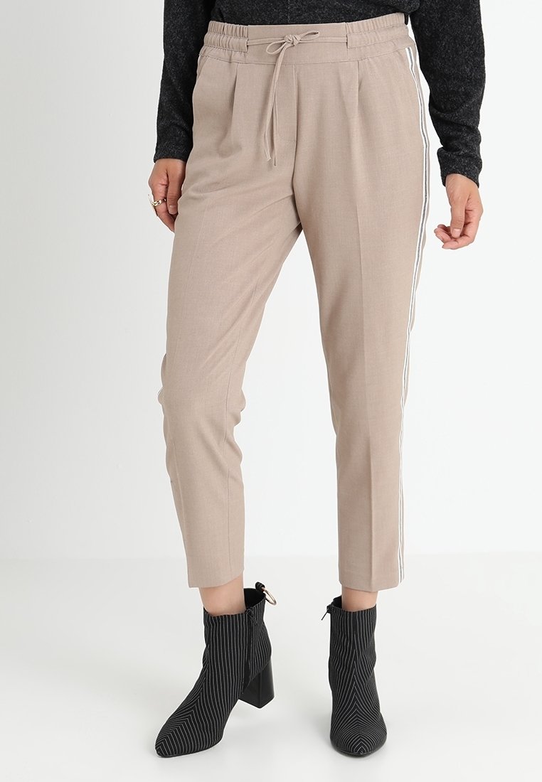 Pantalon beige fuselé avec des bandes blanches sur les côtés, doté d'une taille élastique et d'un cordon de serrage. Porté avec des bottines noires à rayures.