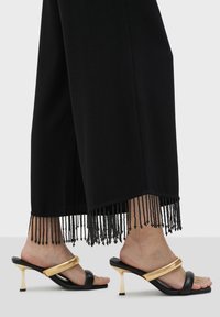 Svarta vida byxor med svarta fransdetaljer i nederkant, parat med svarta och guld klacksandaler som har en böjd guldaccent.