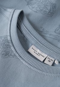 Jasnoniebieska bluza z krótkim rękawem i prążkowanym, okrągłym dekoltem. Zawiera subtelne rysunki liniowe oraz metkę z napisem "basic needs by name it."