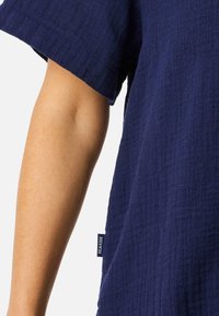 Chemise en coton bleu marine, à tissage lâche et à coupe décontractée. Dotée de manches courtes et d'une petite étiquette "SEASIDE" sur la couture latérale.